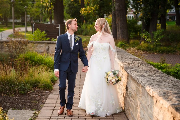 Bride and groom walk together along the stone wall outside Lake Ellyn Boathouse in Glen Ellyn, IL